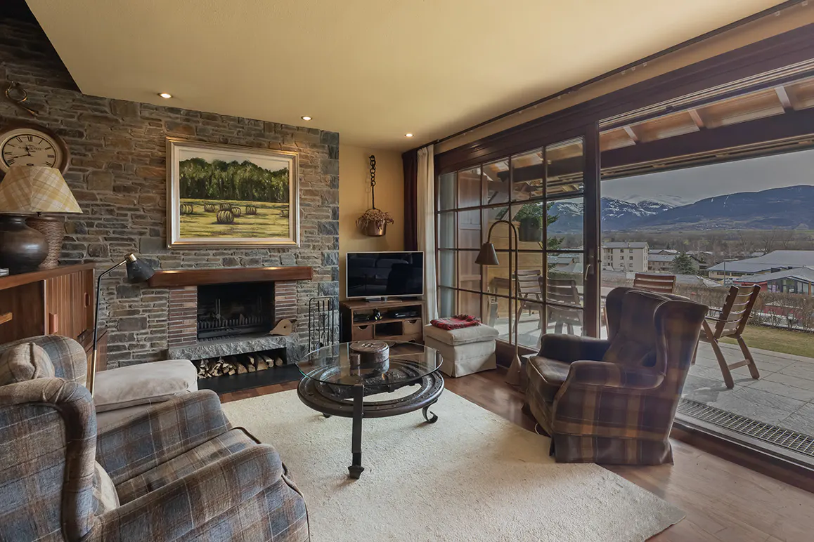 Living room with stone fireplace, plaid armchairs, and glass coffee table. Sliding glass doors open to a patio with mountain views.