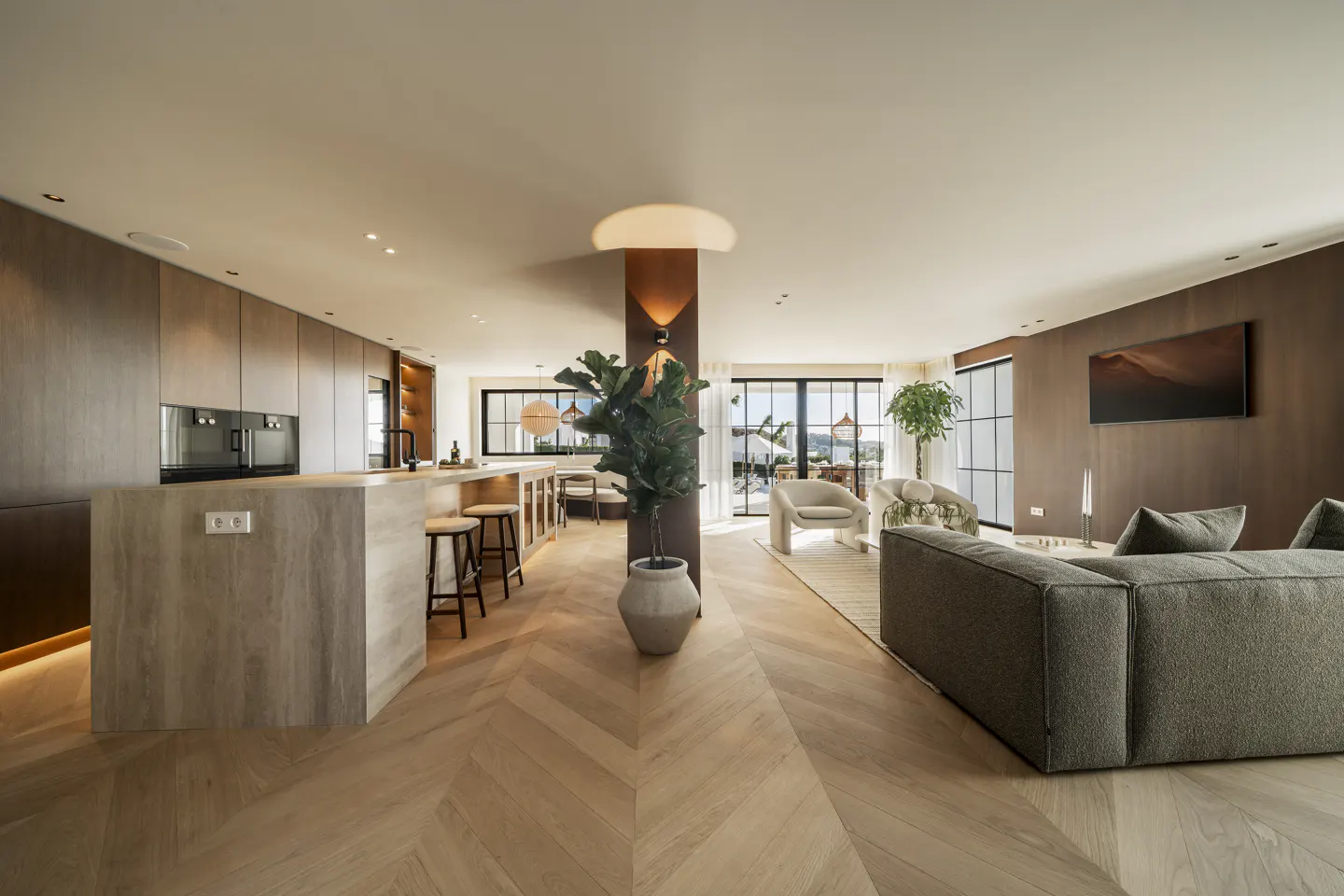 Open-concept living space with wood floors, a kitchen island with stools, and a gray sofa. Large windows offer natural light.