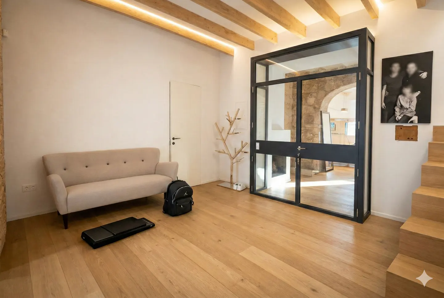 Bright foyer with wood floors, a beige sofa, and a black-framed glass door. A wooden coat rack and stairs are visible. Beams line the ceiling.