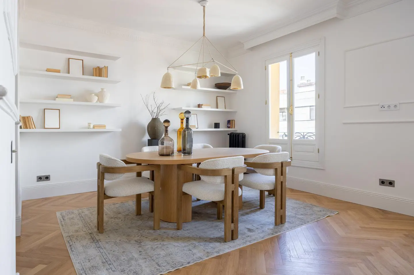 Bright dining room with a round wooden table, six chairs, and a patterned rug. Shelves line the wall, and a modern chandelier hangs above.
