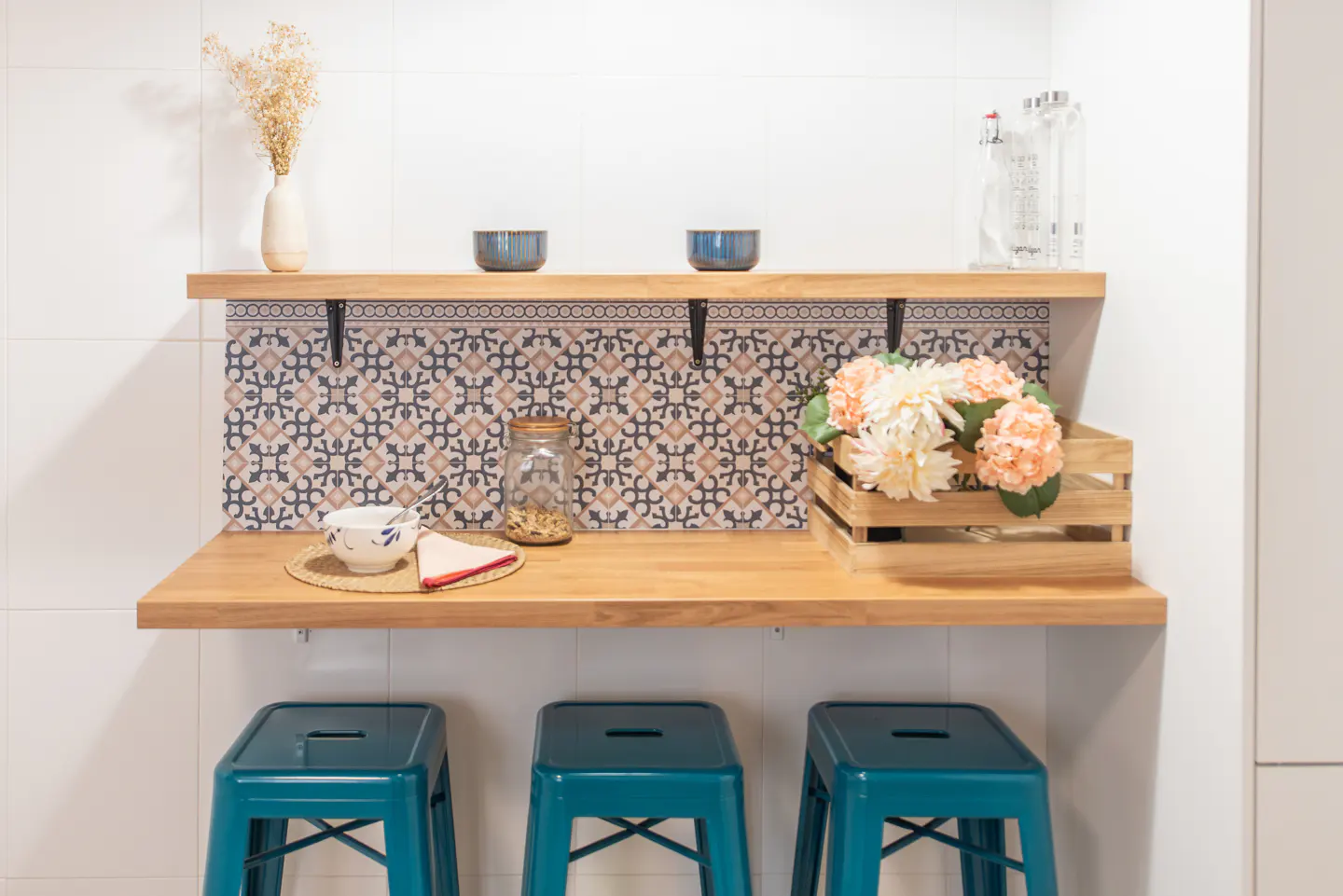 A kitchen counter with teal stools, a wooden shelf, and decorative items. The backsplash has a patterned tile design.