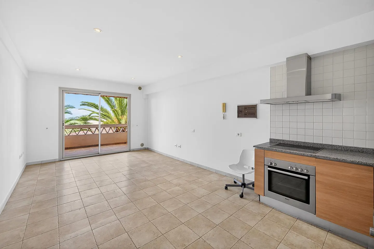 Bright, empty apartment with tile floors, white walls, and a sliding glass door to a balcony with palm trees. A kitchen area features wood cabinets and stainless steel appliances.