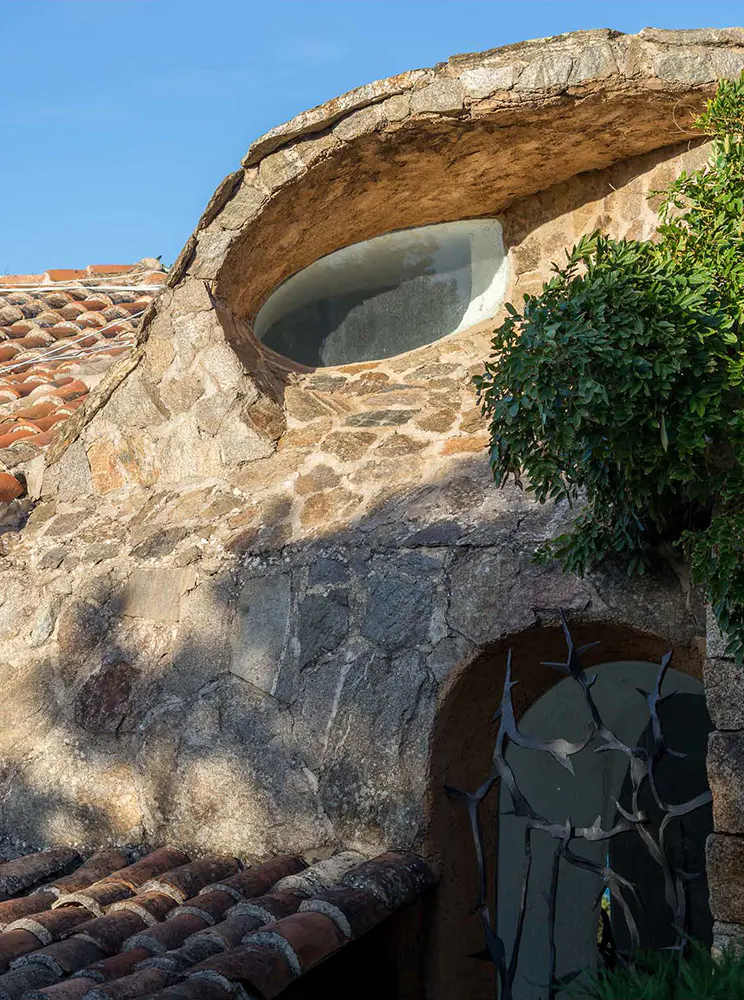 Close-up of a stone building with a round window and a red tile roof. Green foliage is visible on the right side of the image.