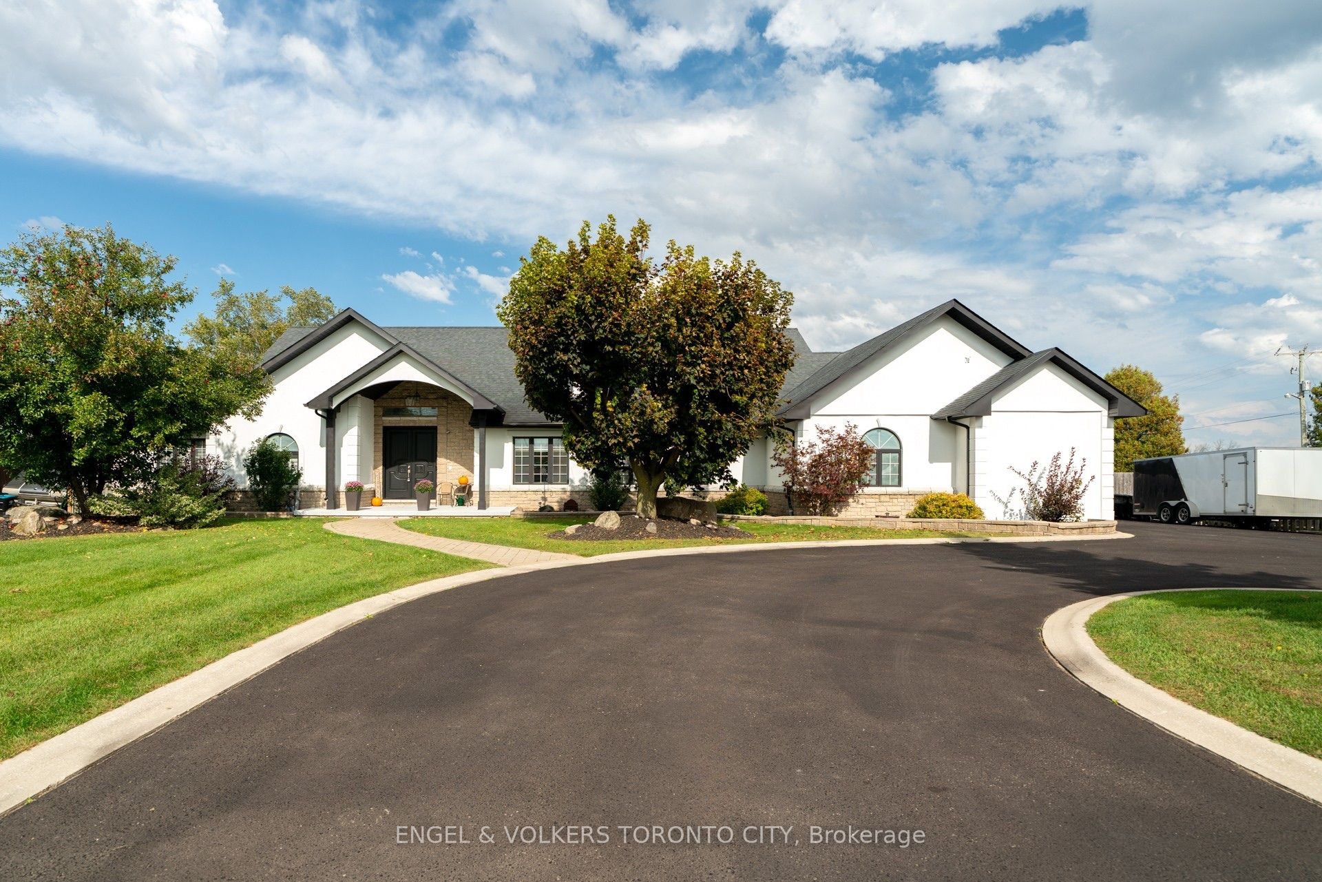 White single-story house with a dark roof, curved asphalt driveway, and green lawn under a blue sky with scattered clouds. [AI-generated text]