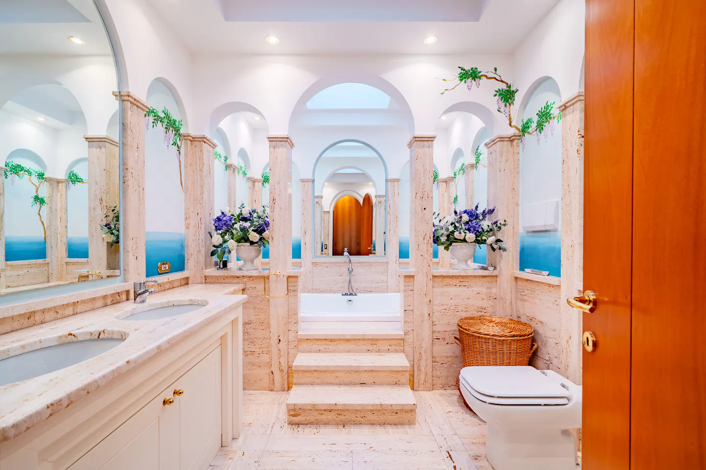 Elegant bathroom with marble columns, arches, and a mural of the sea. A white tub sits on a raised platform with steps. A toilet and vanity are also visible.