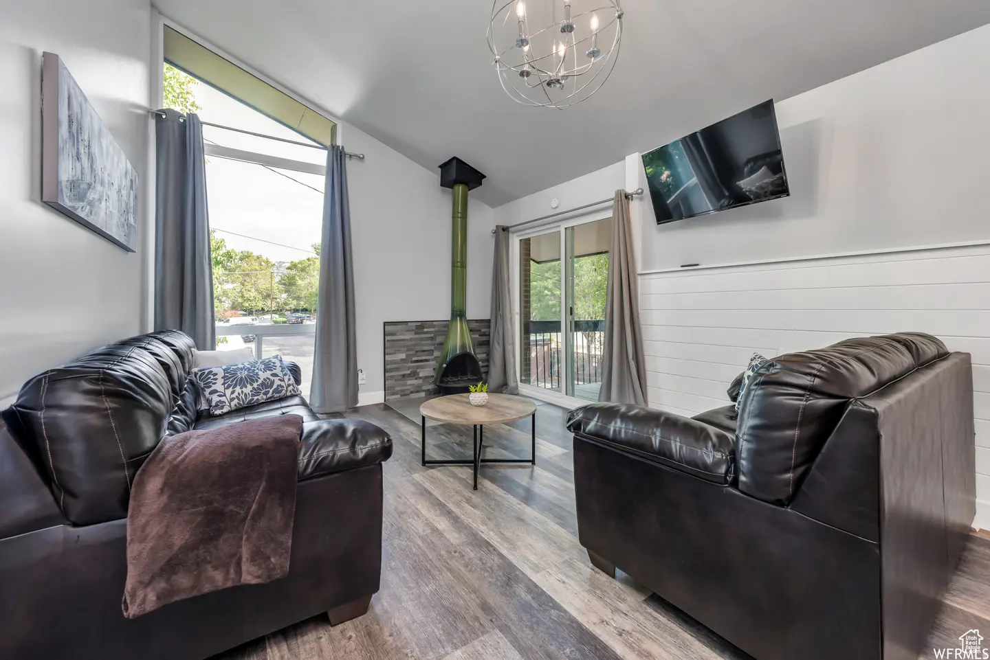 Living room with gray wood floors, black leather sofas, and a green fireplace. A round table sits in the center. Curtains frame the windows.