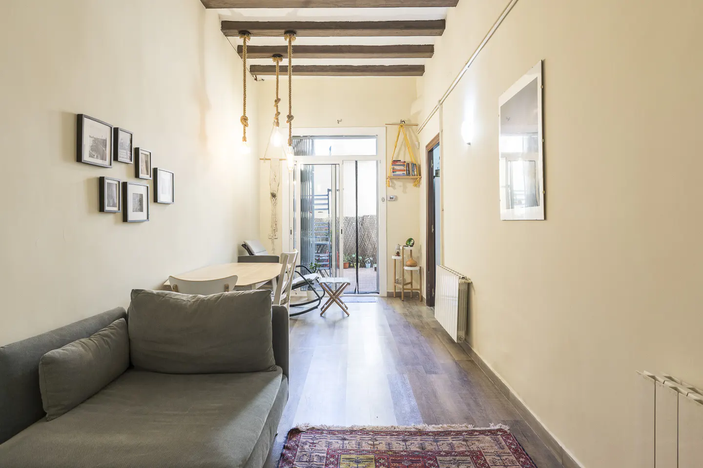 A long, narrow living room with a gray sofa, dining table, and balcony. The walls are cream-colored, and the floor is wood. There is a rug on the floor.