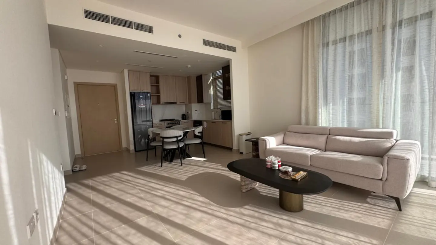 Modern open-plan apartment with a beige sofa, oval coffee table, dining area, and kitchen with light wood cabinets.