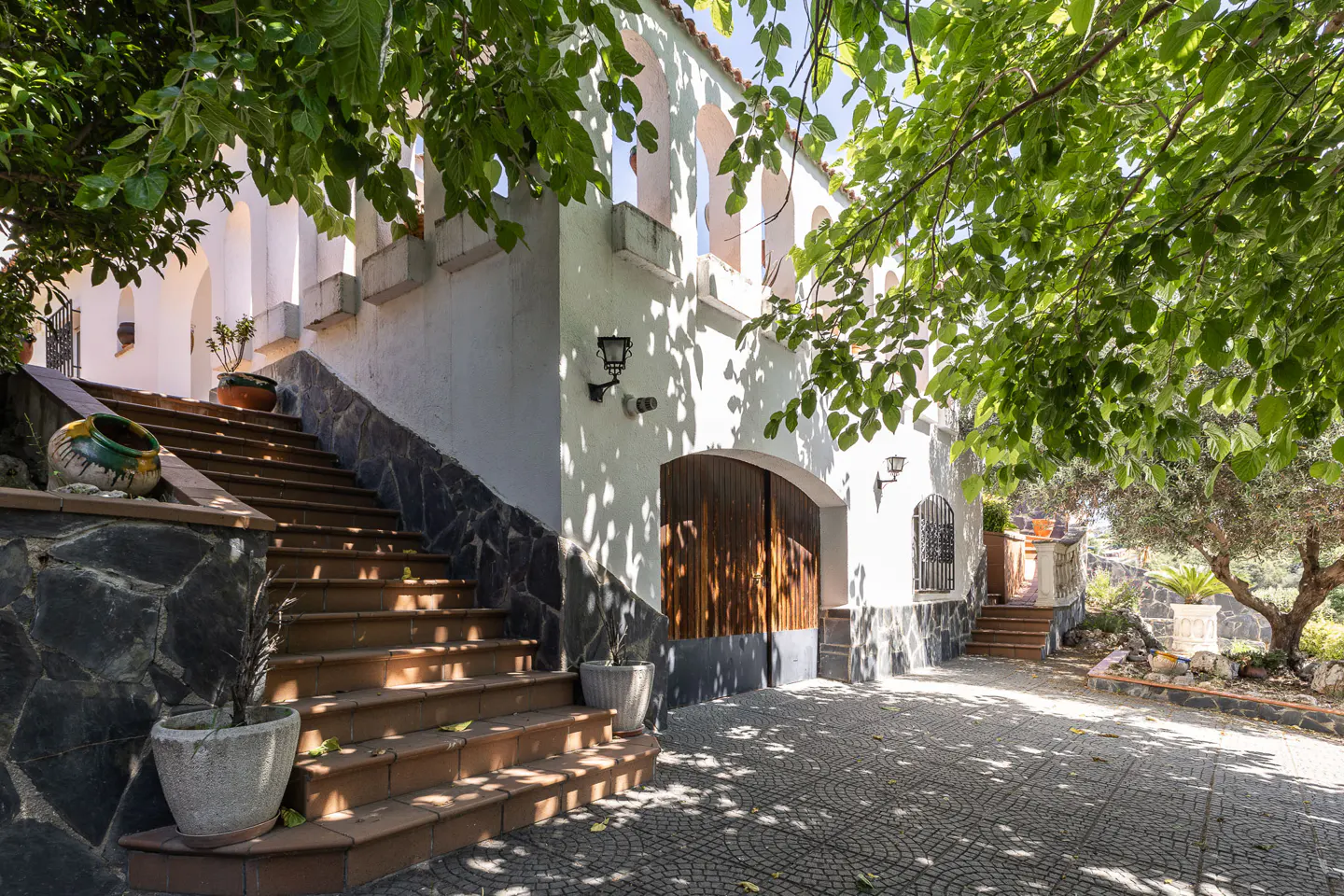 White villa exterior with arched balcony, stone-tiled stairs, wooden garage doors, and lush trees in a sunny courtyard.