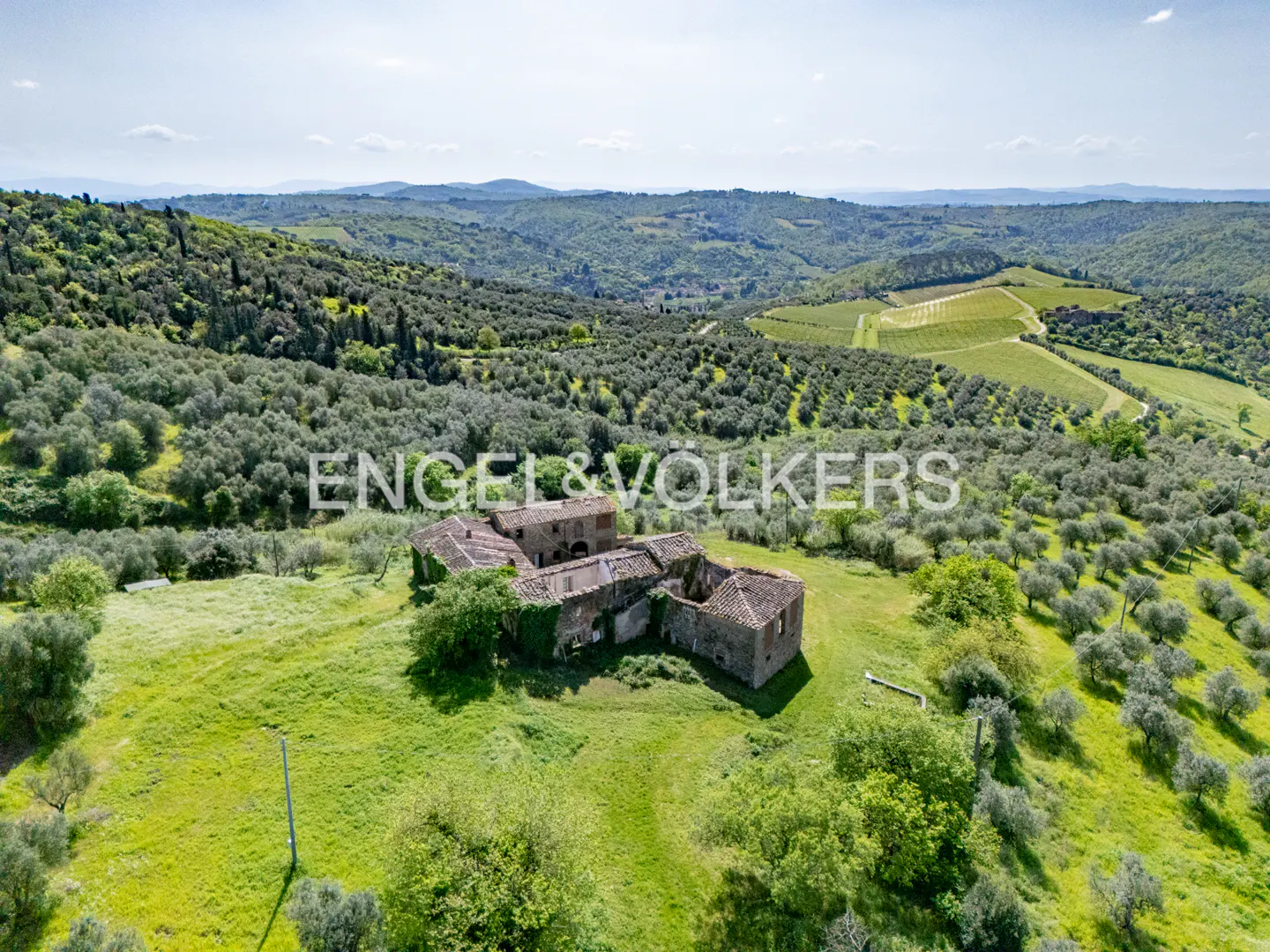 Aerial view of a stone farmhouse with a red tile roof, surrounded by green hills and olive trees in Tuscany.