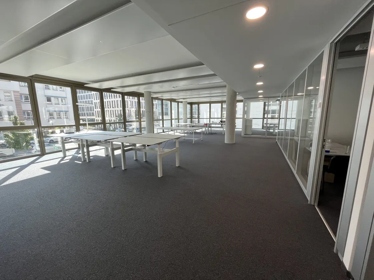 Bright, empty office space with gray carpet, white desks, and large windows showing a city view. Glass walls divide the space.