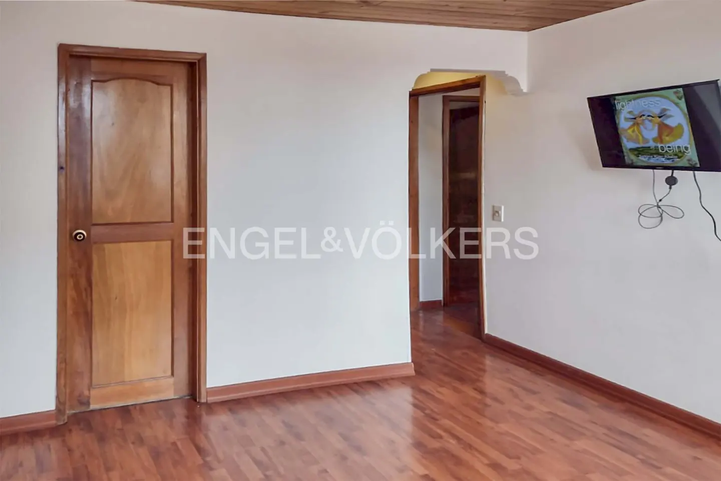 A room with wood floors, white walls, and a wood ceiling. A TV hangs on the wall. Two wood doors are visible.