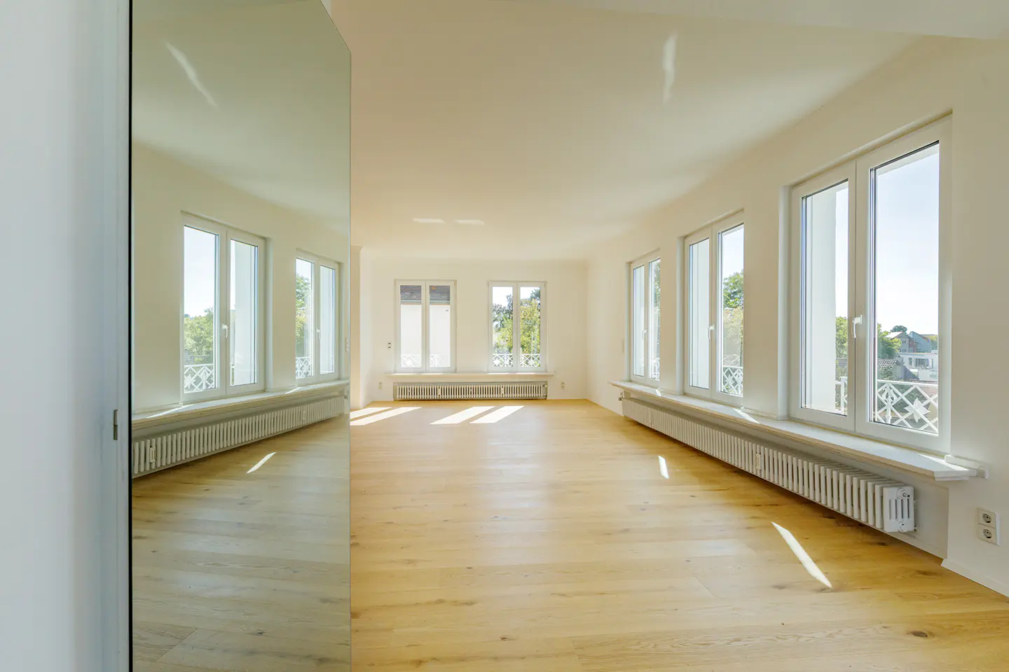Bright, empty room with light wood floors, white walls, and multiple windows. A large mirror reflects the room. Radiators are under the windows.
