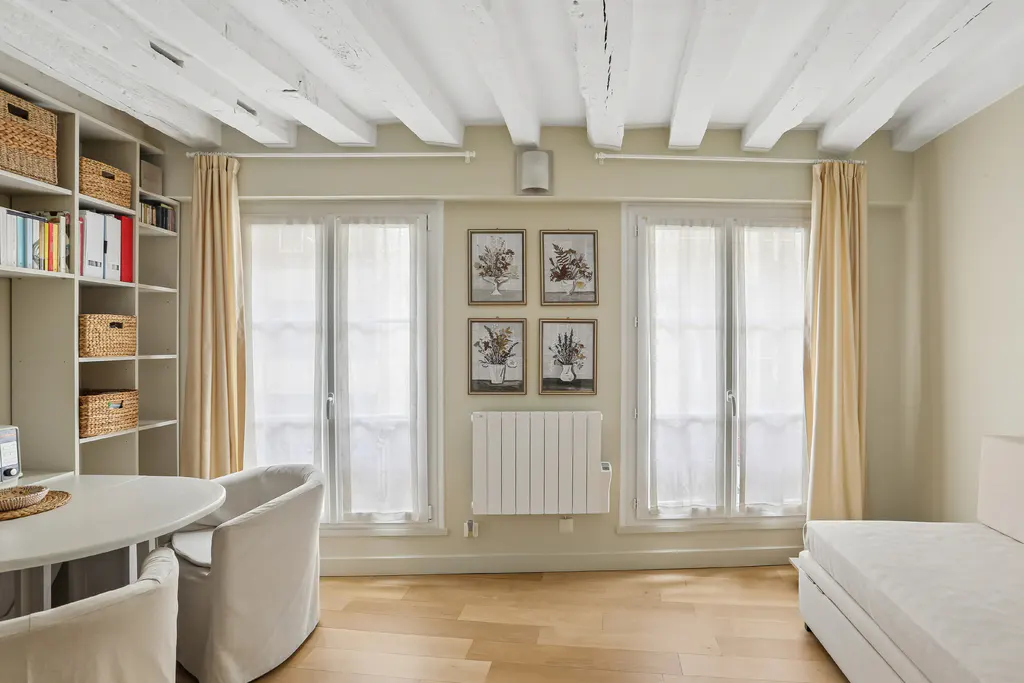 Bright room with white wood-beamed ceiling, two windows with beige curtains, and a white bookshelf with wicker baskets. Four floral paintings hang above a white radiator. A white table and chairs are in the foreground.