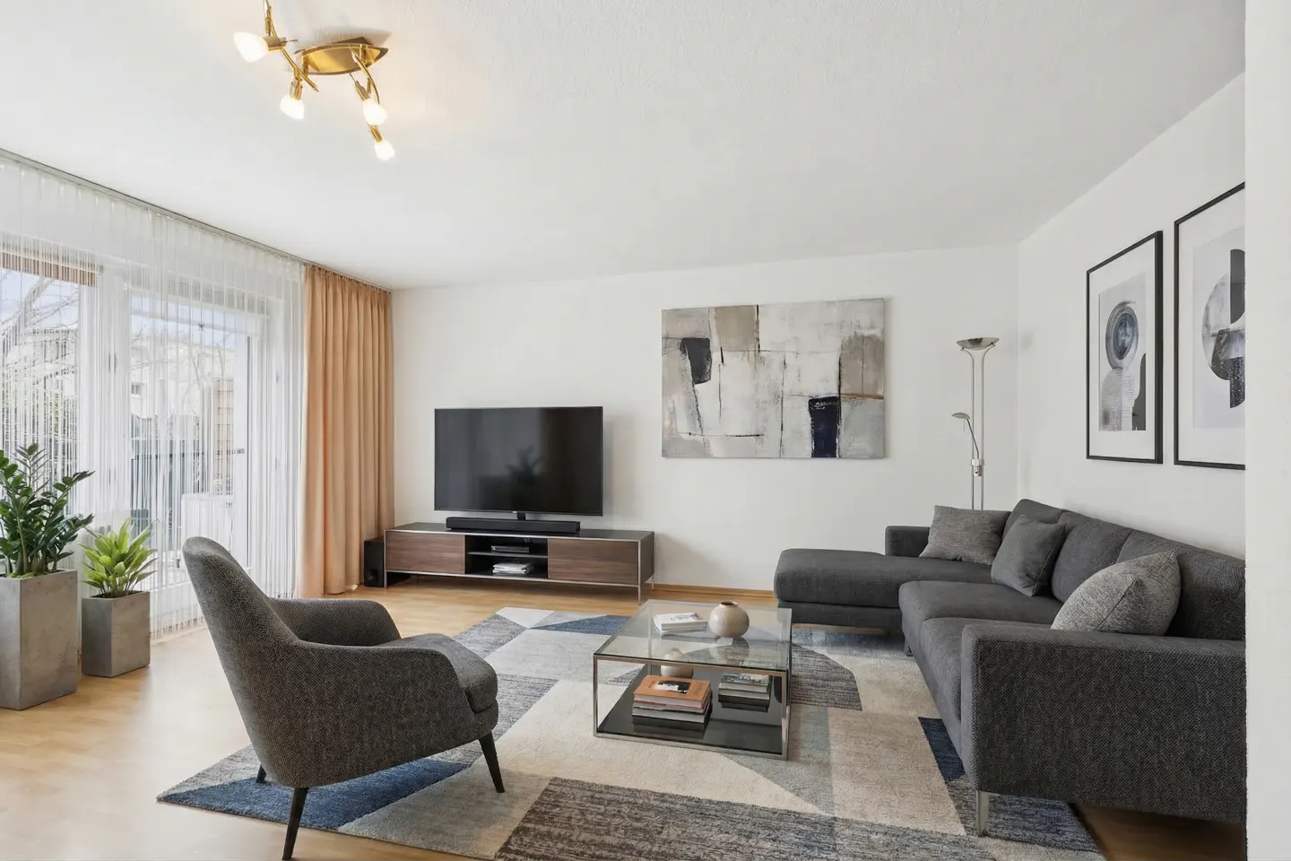 Bright living room with gray sectional sofa, armchair, and geometric rug. A TV sits on a wooden console table. Artwork adorns the white walls.