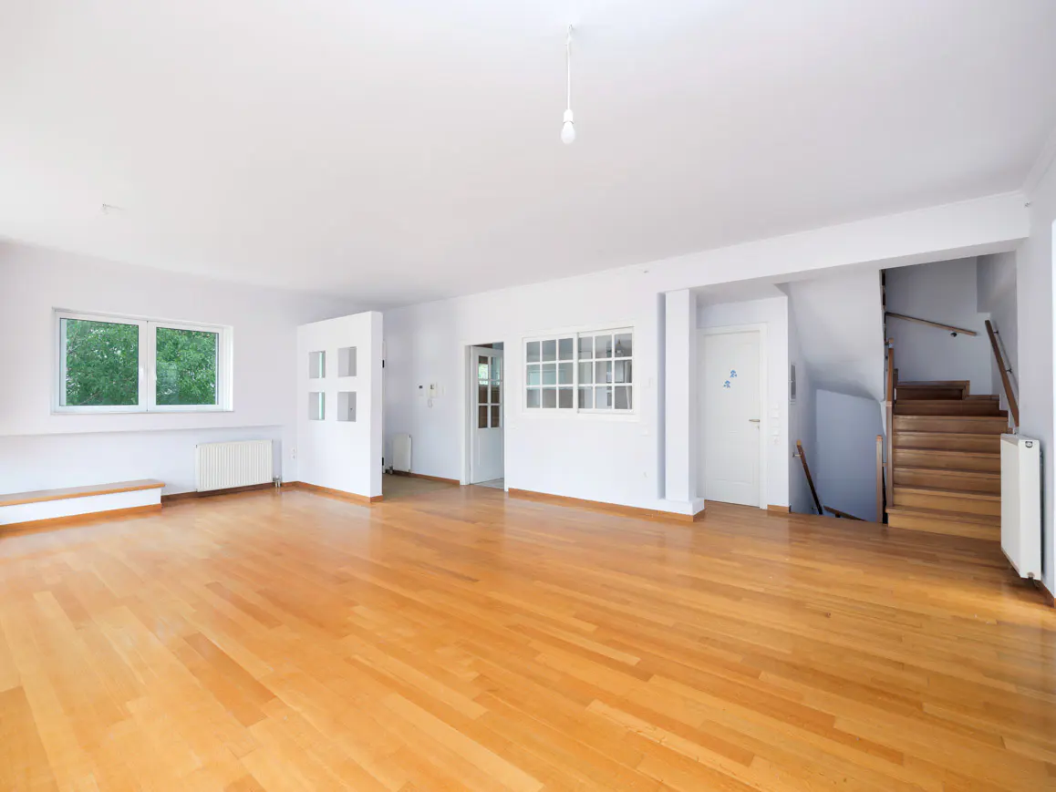 Bright, empty room with hardwood floors, white walls, windows, and a staircase. A single lightbulb hangs from the ceiling.