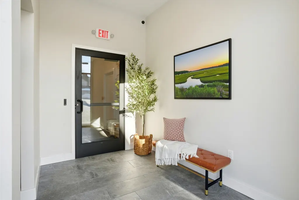 Entryway with gray tile floor, black door with "EXIT" sign, bench with pillow and blanket, plant, and landscape art.