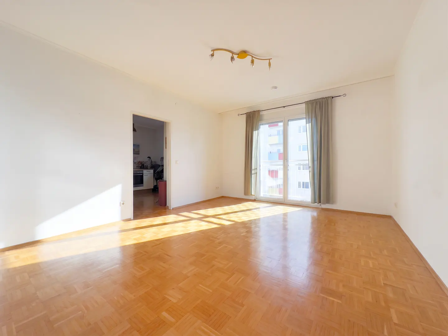 Bright, empty room with parquet floor, white walls, and a door to the kitchen. Sunlight streams in from a window with gray curtains.