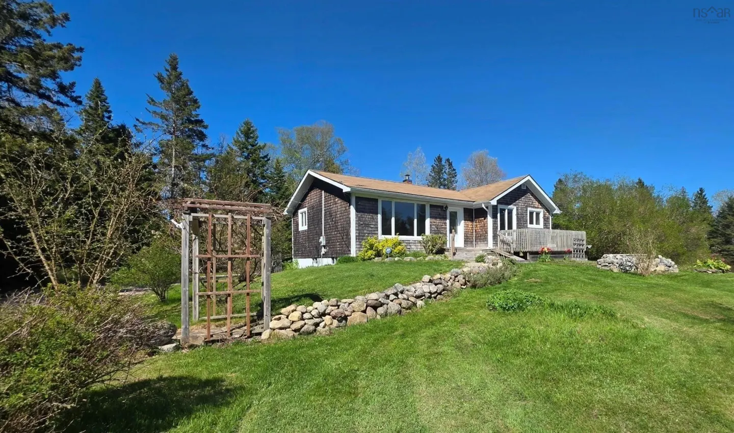 A shingled cottage sits on a grassy hill with a stone retaining wall, a wooden trellis, and a clear blue sky background.