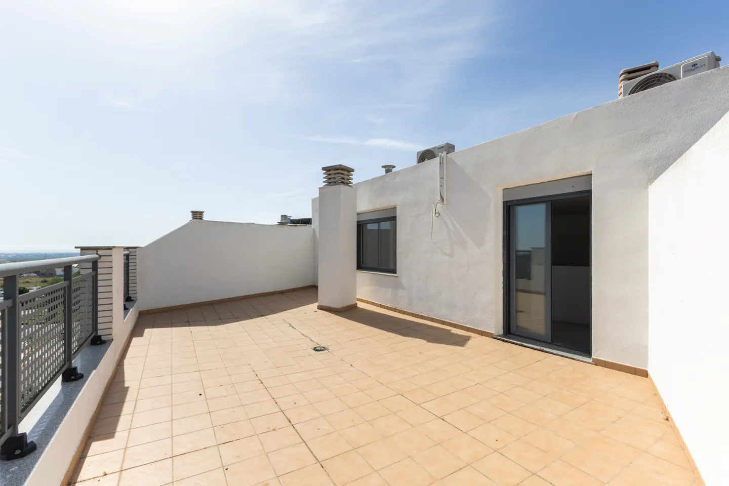 A rooftop terrace with beige tile flooring, white walls, and a metal railing overlooking a distant landscape under a blue sky.