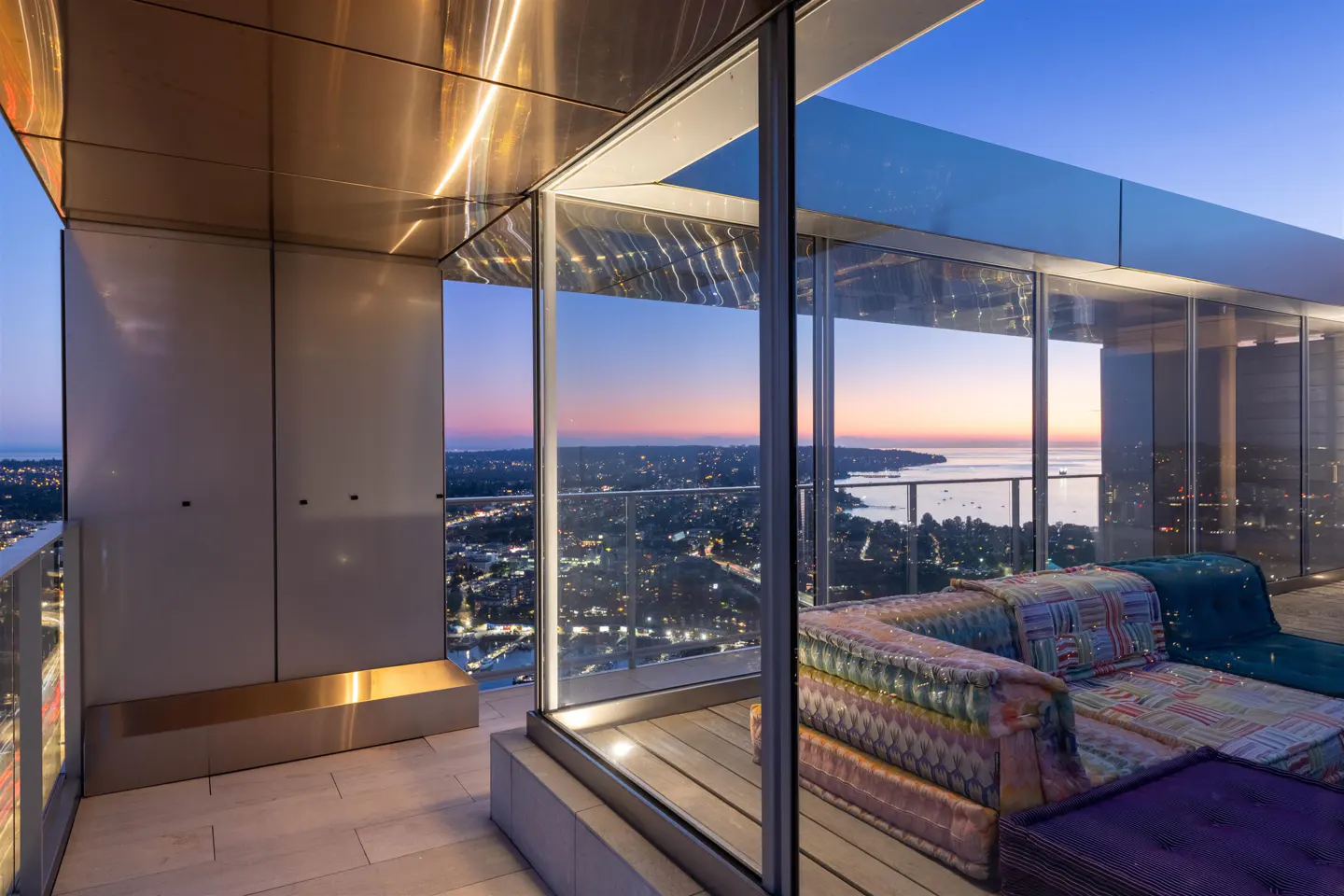 Modern condo balcony with a colorful couch and a city view at dusk. The balcony has glass walls and a metal ceiling.