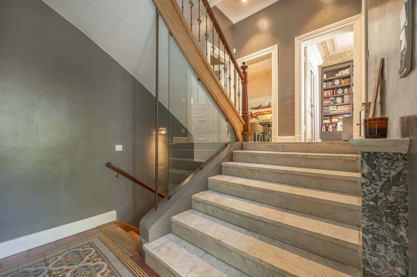 Interior view of a staircase with marble steps, a wooden handrail, and a glass panel, leading to doorways and a bookshelf.