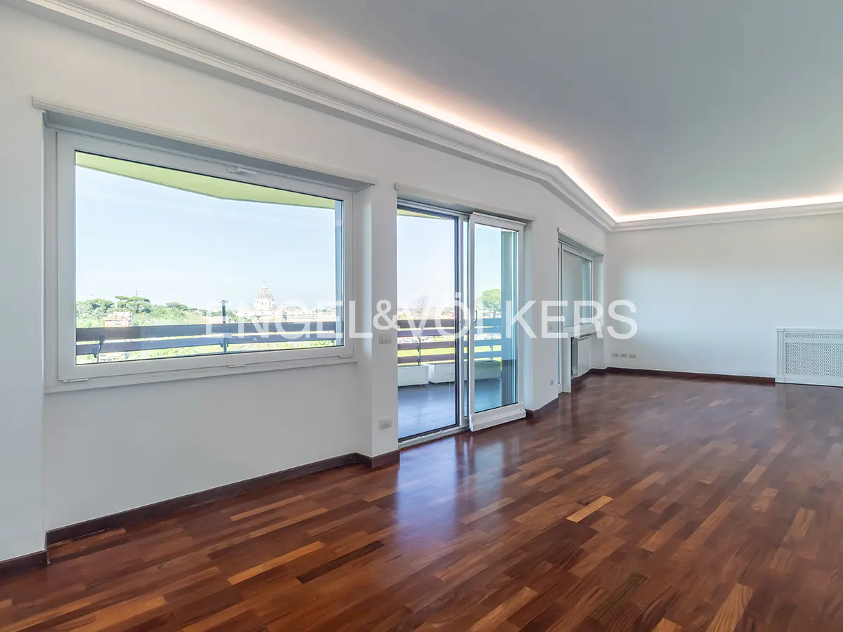 Bright, empty room with hardwood floors, white walls, and large windows with a city view. Sliding glass doors lead to a balcony.