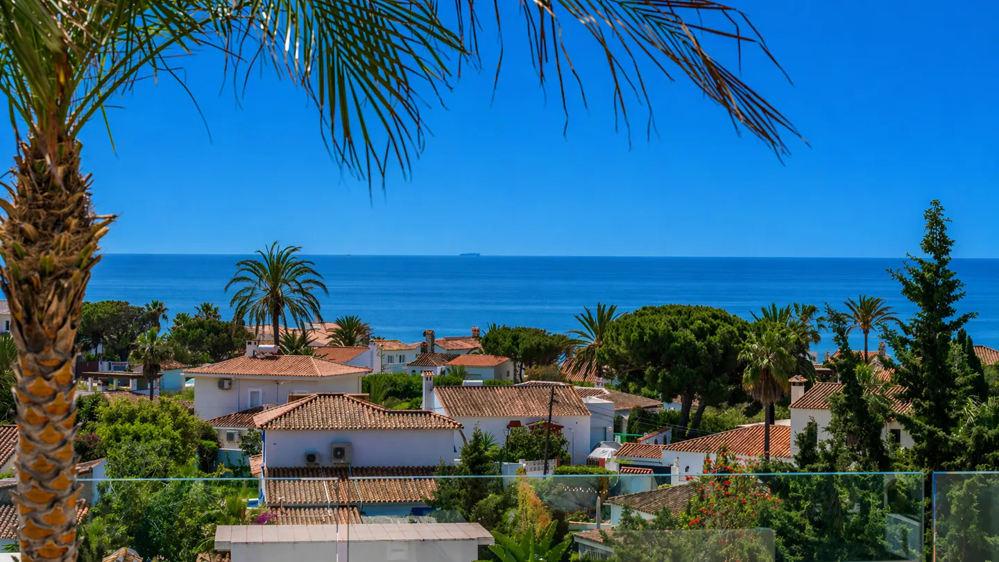 View of white houses with orange tile roofs, green trees, and blue ocean under a clear sky. Palm tree in foreground.