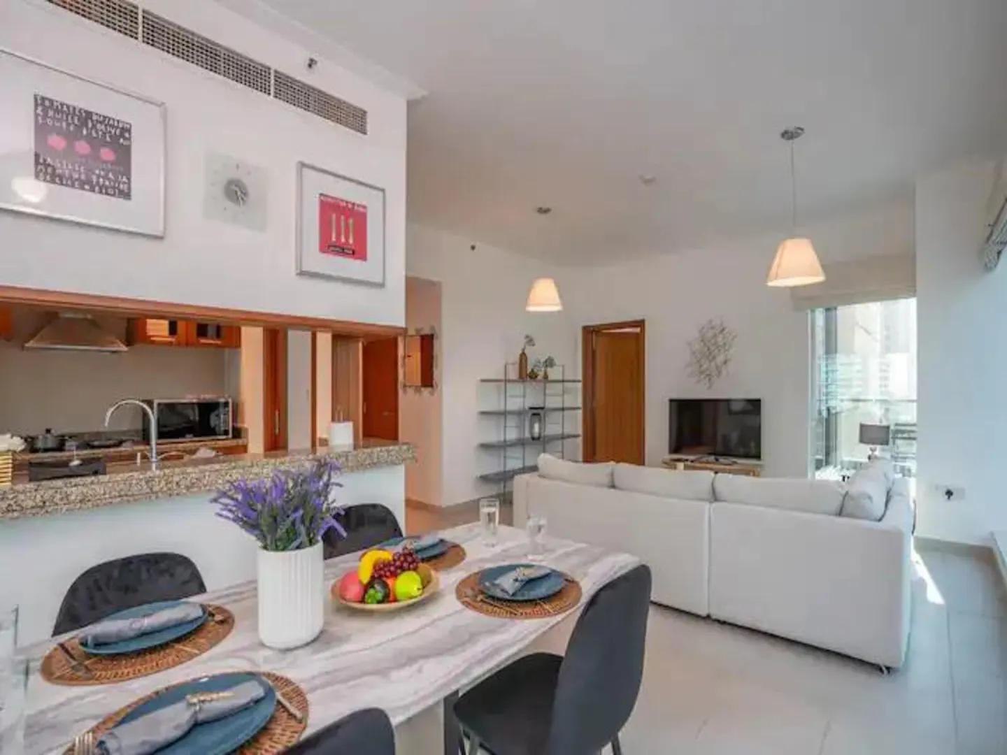 Modern open-plan apartment with a marble dining table, fruit bowl, and a white sectional sofa in the living area.