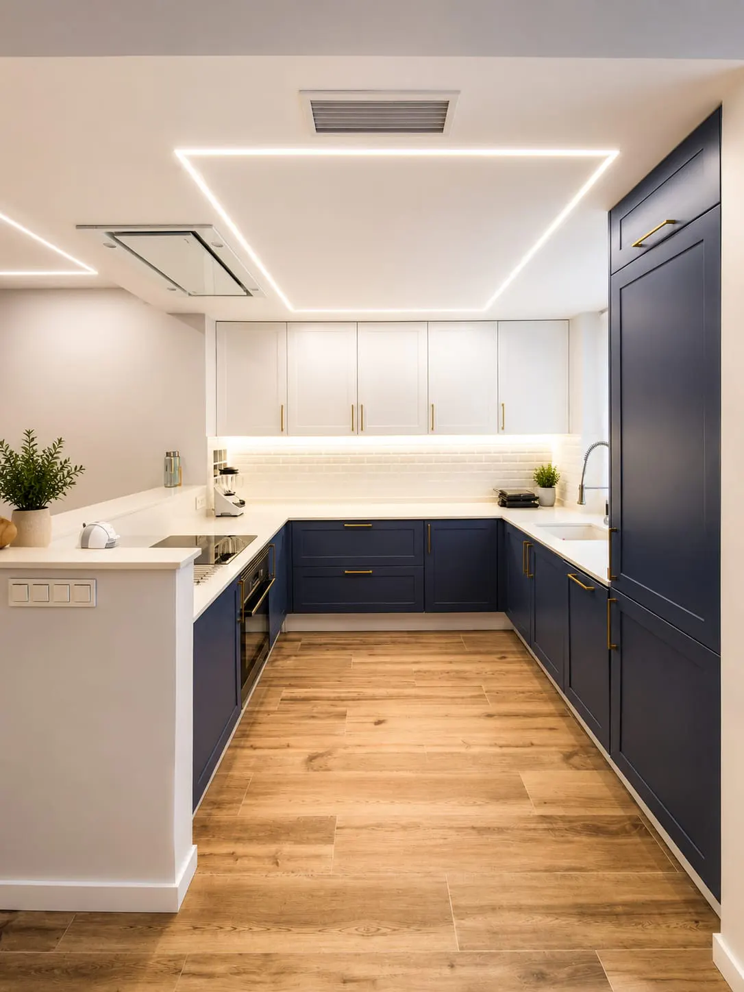 A modern kitchen with white and navy blue cabinets, gold handles, and wood-look tile flooring. The ceiling has recessed lighting and a square LED light fixture.