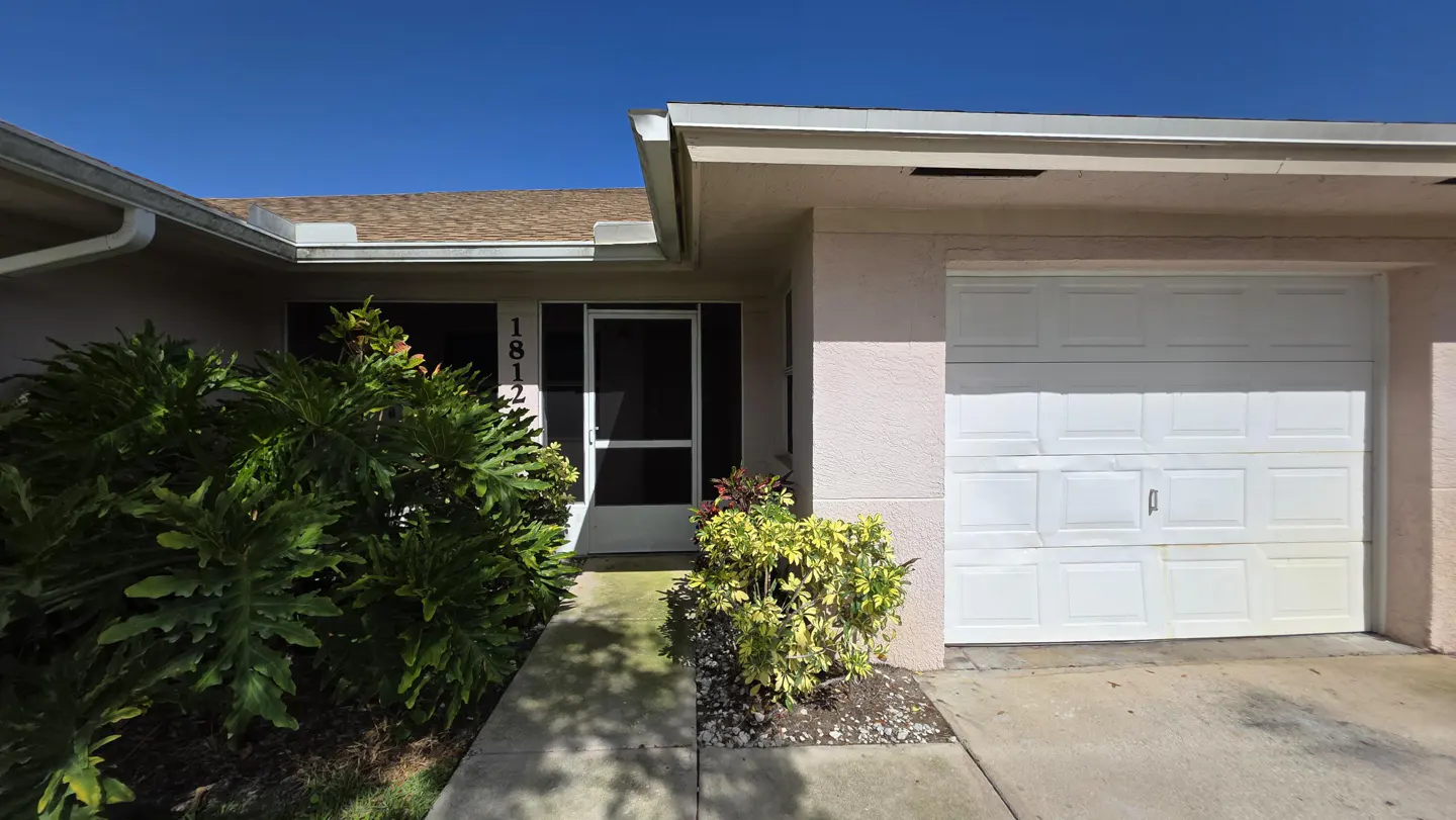 Exterior of a one-story home with a white garage door, screen door, and lush green plants. The house number "1812" is visible.