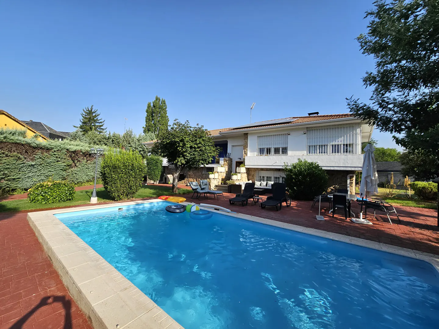 A backyard with a blue swimming pool, lounge chairs, and a white two-story house in the background on a sunny day.