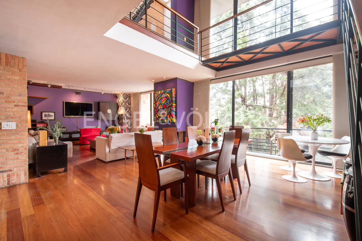 Open-concept living and dining area with hardwood floors, purple walls, and floor-to-ceiling windows overlooking trees.