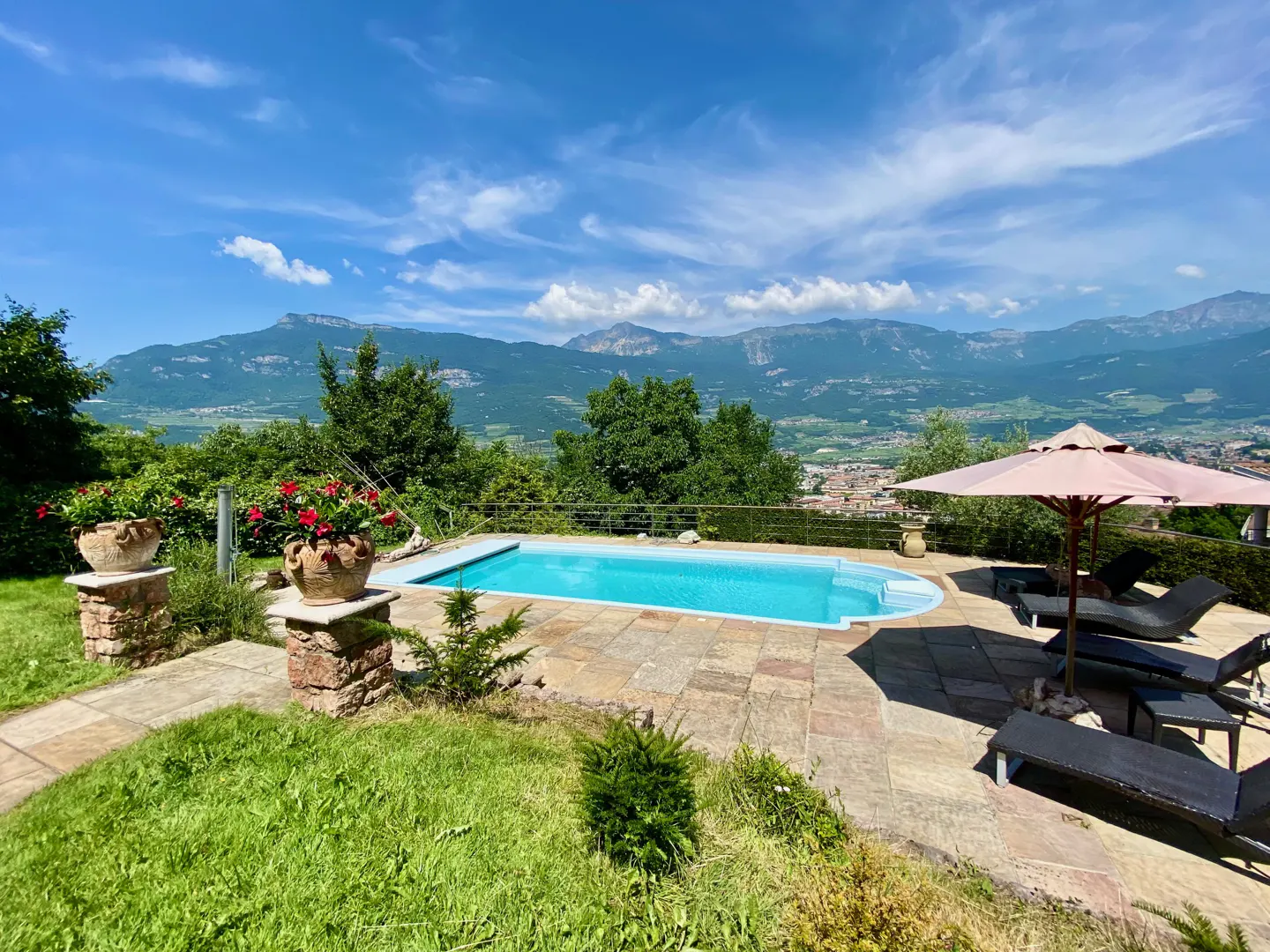 Outdoor pool with lounge chairs and umbrella, stone patio, green lawn, trees, and mountain view under a blue sky.