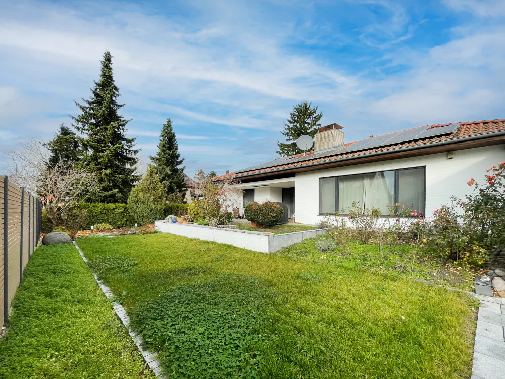 A white house with a red tile roof and solar panels, surrounded by a green lawn and trees under a blue sky.