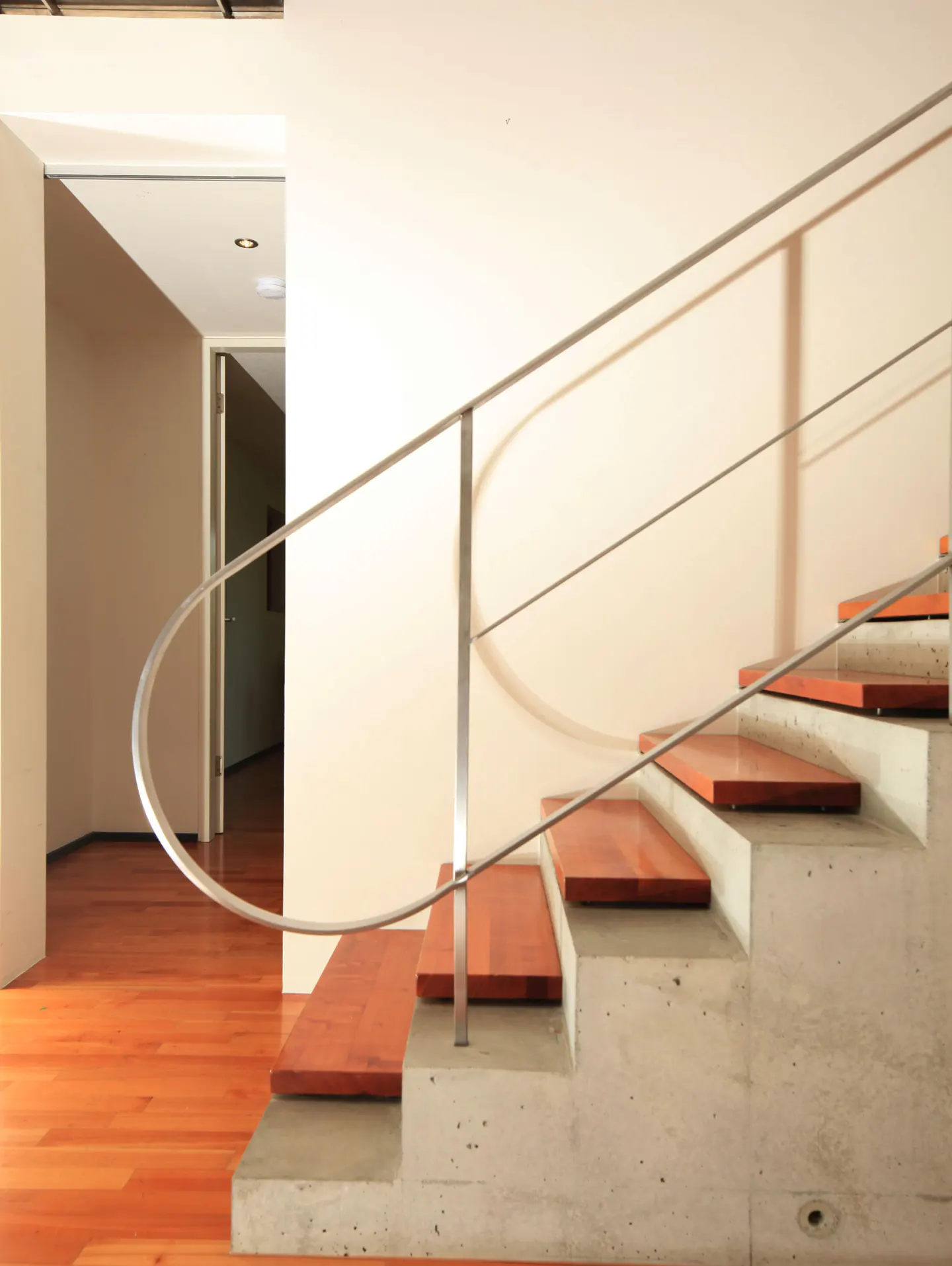 Interior view of modern staircase with wood treads, concrete risers, and a curved metal handrail against a light beige wall.