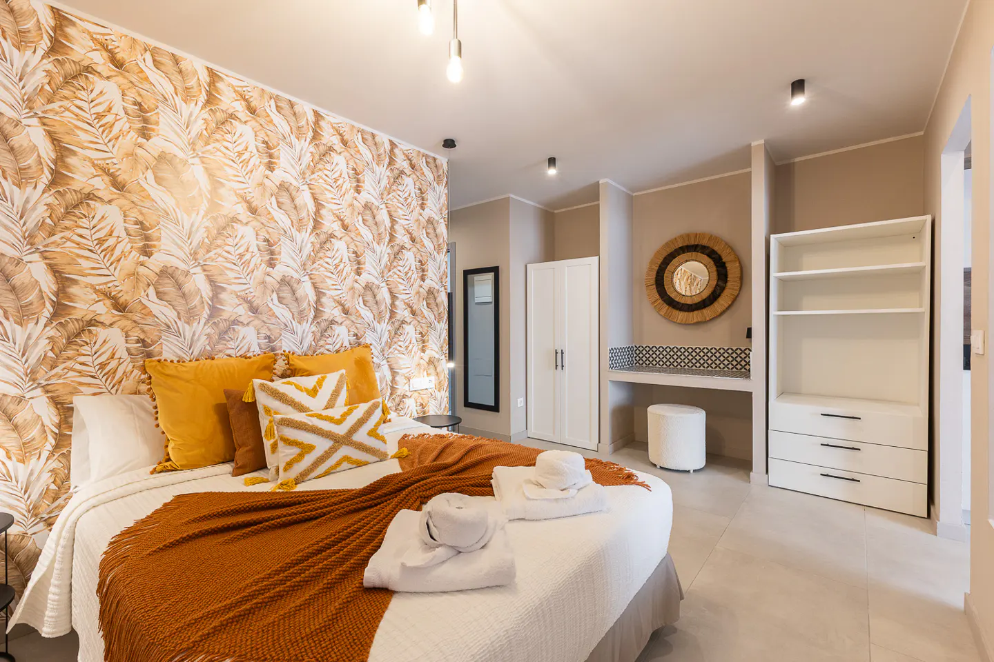Bedroom with leaf-patterned wallpaper, white bed with orange blanket and pillows, and white closet and shelves.