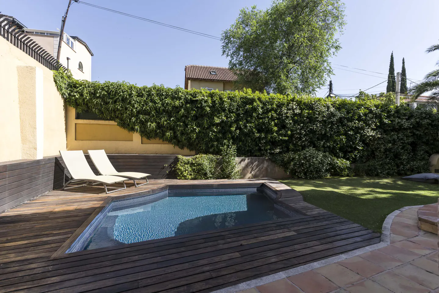 A backyard pool with blue water is surrounded by a dark wood deck and two lounge chairs. A green hedge provides privacy.