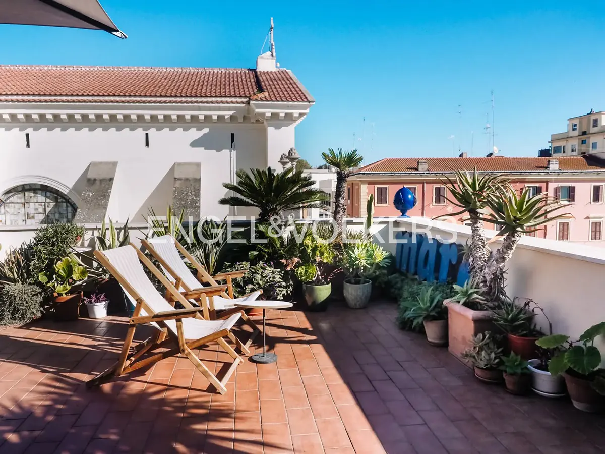 Rooftop terrace with two white lounge chairs, plants in pots, and a red brick floor. White building and blue sky in the background.