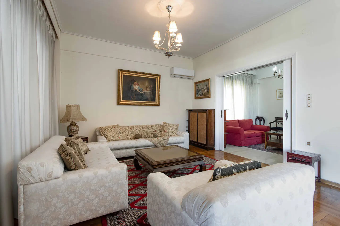 A living room with white walls, patterned sofas, a chandelier, and a red patterned rug. A doorway leads to another room with a red sofa.