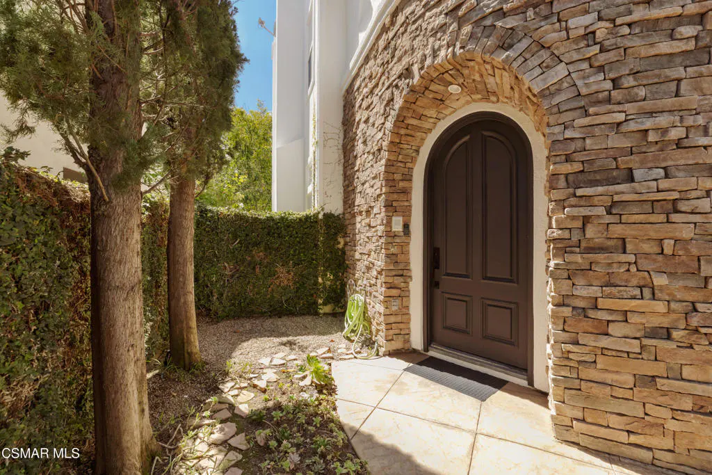 Arched brown front door with stone surround. Trees and green hedge line the walkway to the entrance.