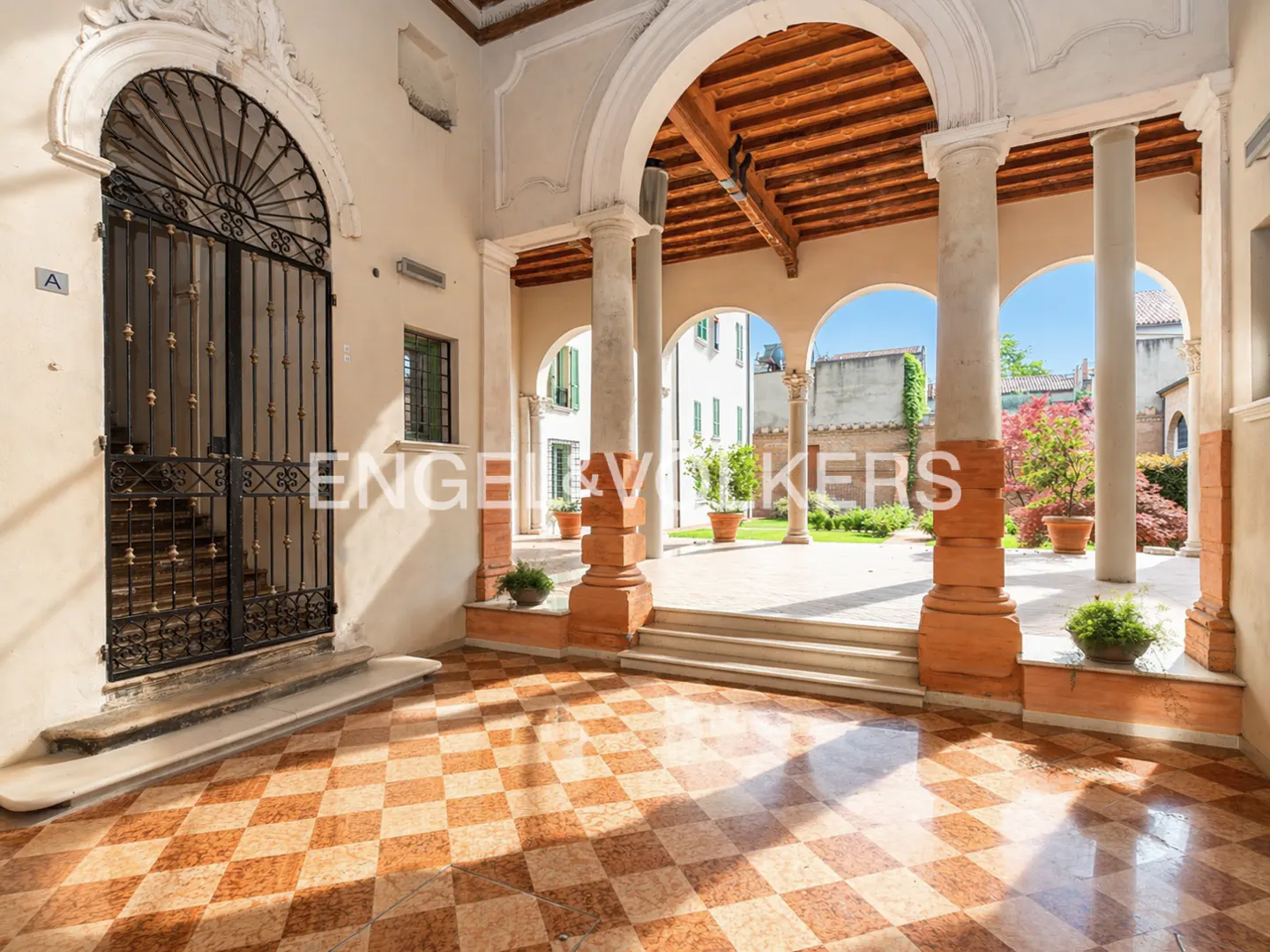 Courtyard with checkered tile floor, arched columns, and wrought iron gate. Potted plants line the walkway leading to a garden.