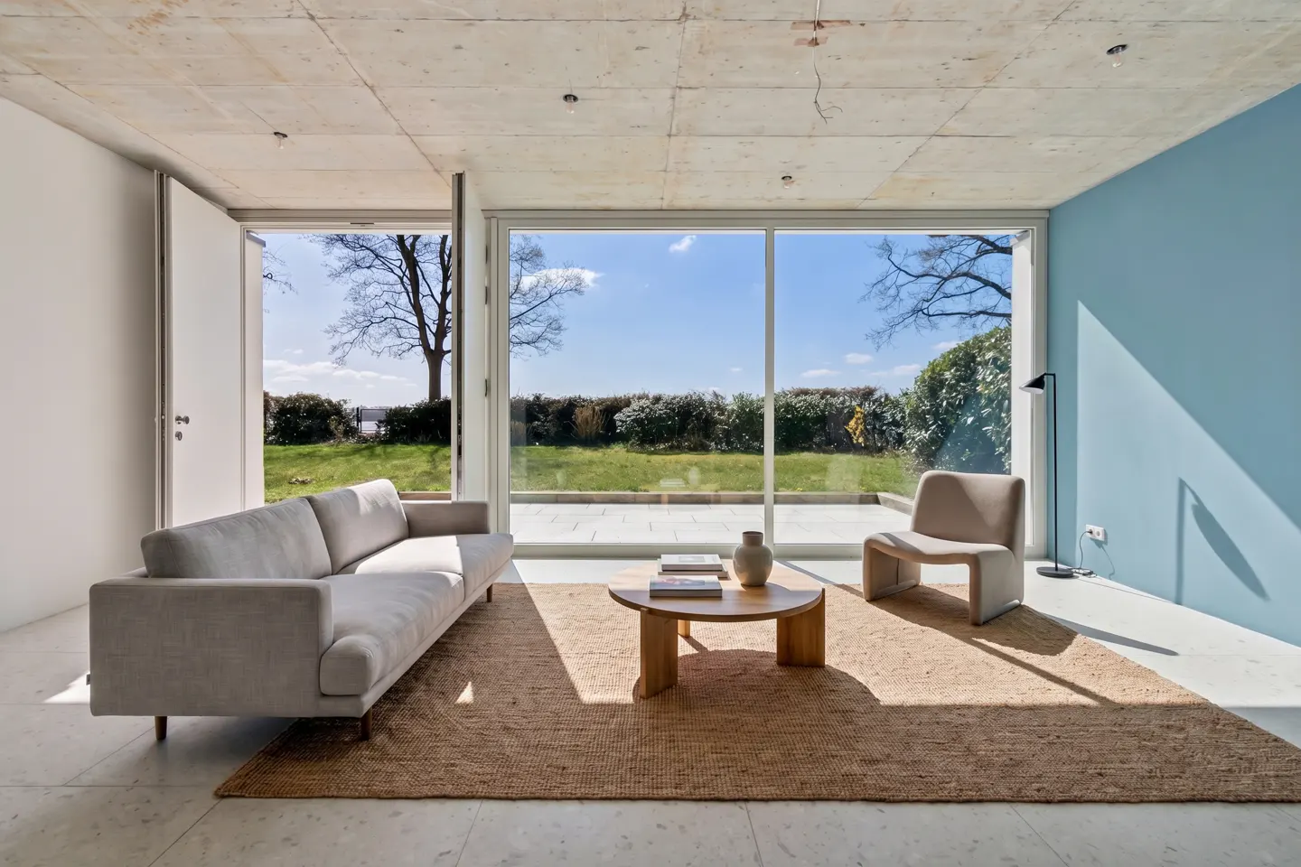 Bright living room with a gray sofa, chair, and round table on a jute rug. Large windows overlook a green lawn and blue sky.
