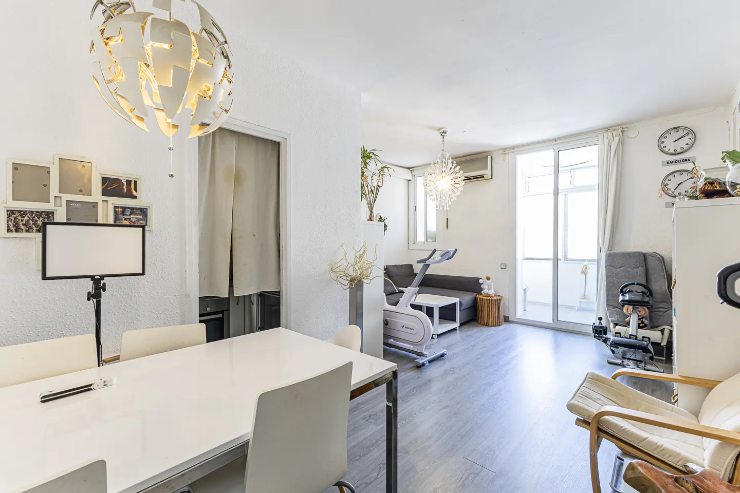 Bright, open-plan apartment interior with white walls, a dining table, and exercise equipment. A modern chandelier hangs above the table.
