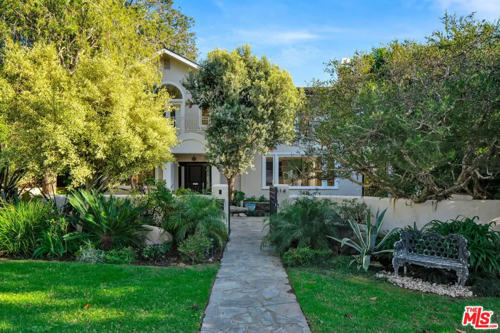 A stone path leads to a two-story light-colored home surrounded by lush trees, green lawns, and a decorative metal bench.