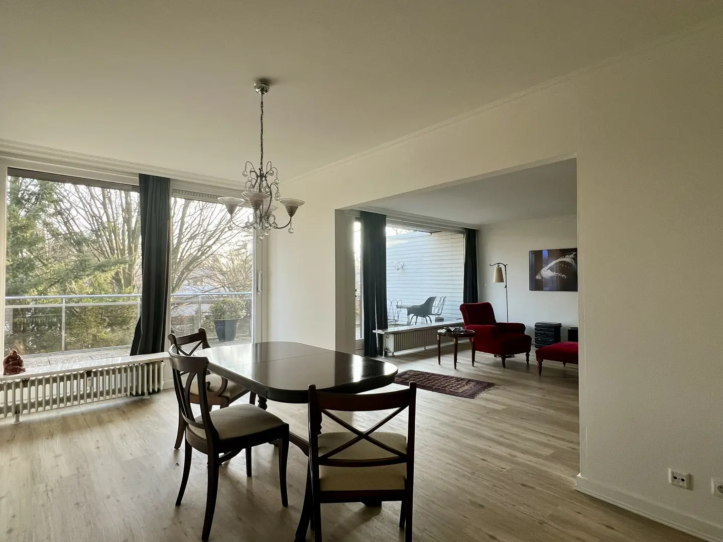 Bright, open-concept dining and living room with wood floors. A dark wood table and chairs sit under a chandelier. Balcony view. Red armchair in living area.