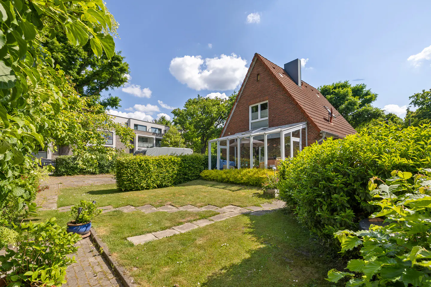 Exterior view of a red brick house with a sunroom, surrounded by green bushes and trees under a blue sky.