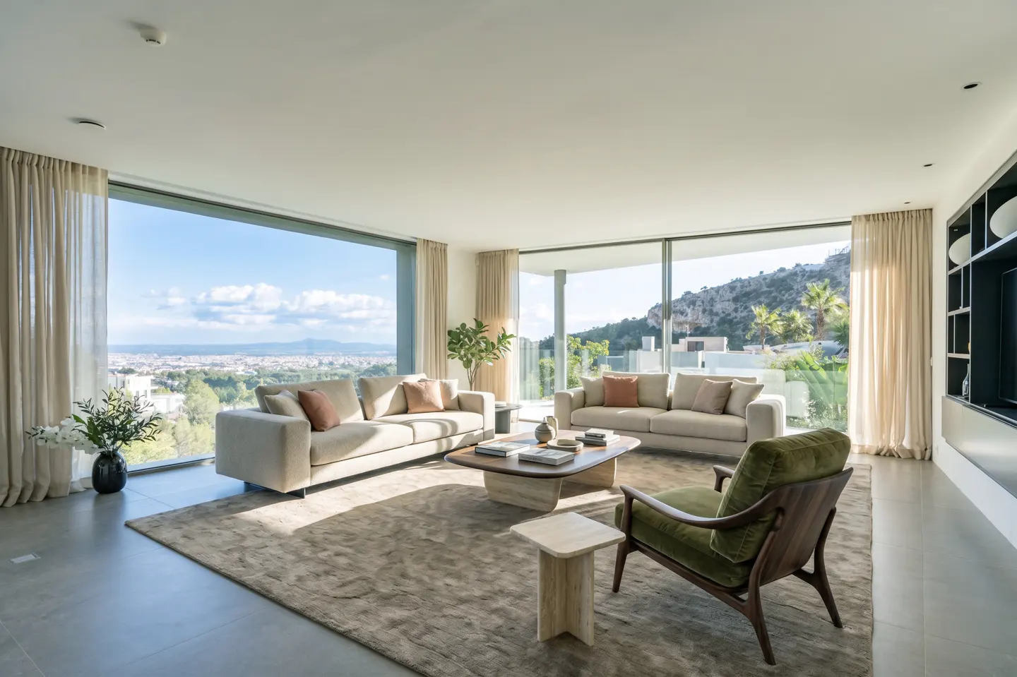 Bright living room with two beige sofas, green armchair, and large windows overlooking a city and mountains.