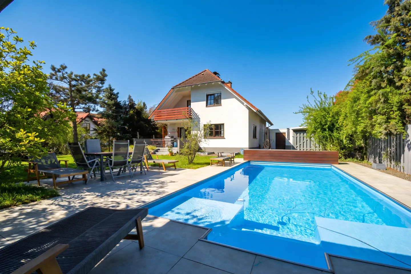 A backyard with a blue swimming pool, a white house with a red roof, and outdoor furniture on a sunny day.