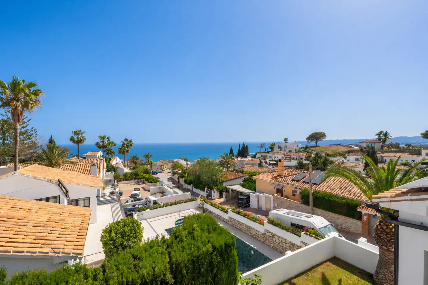 High-angle view of white villas with terracotta roofs, palm trees, and a swimming pool overlooking the blue Mediterranean Sea.